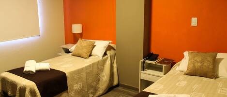 A hotel room with two beds, each with a brown bedspread and white pillows. There is a nightstand with a lamp and a phone on it. The walls are orange, and there are abstract paintings on the walls.