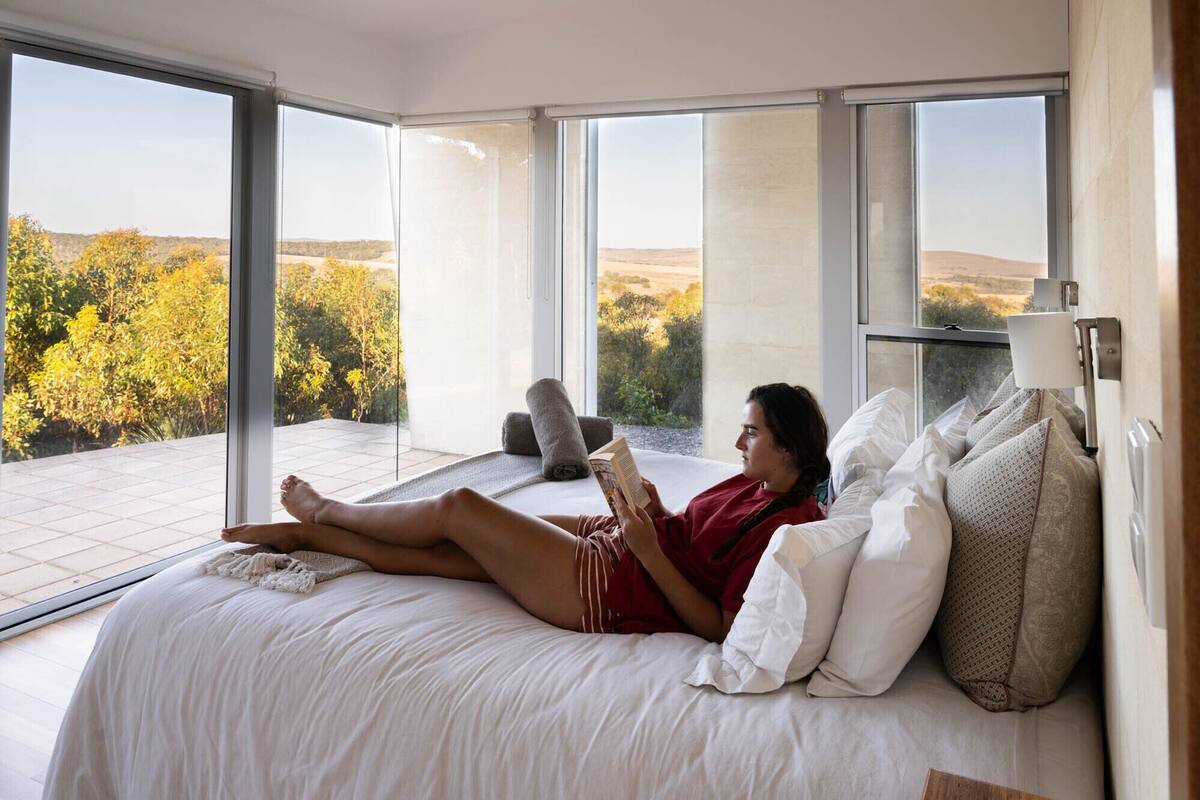 Un dormitorio con una cama grande, varios almohadones y vistas a un paisaje natural a través de amplias ventanas.
