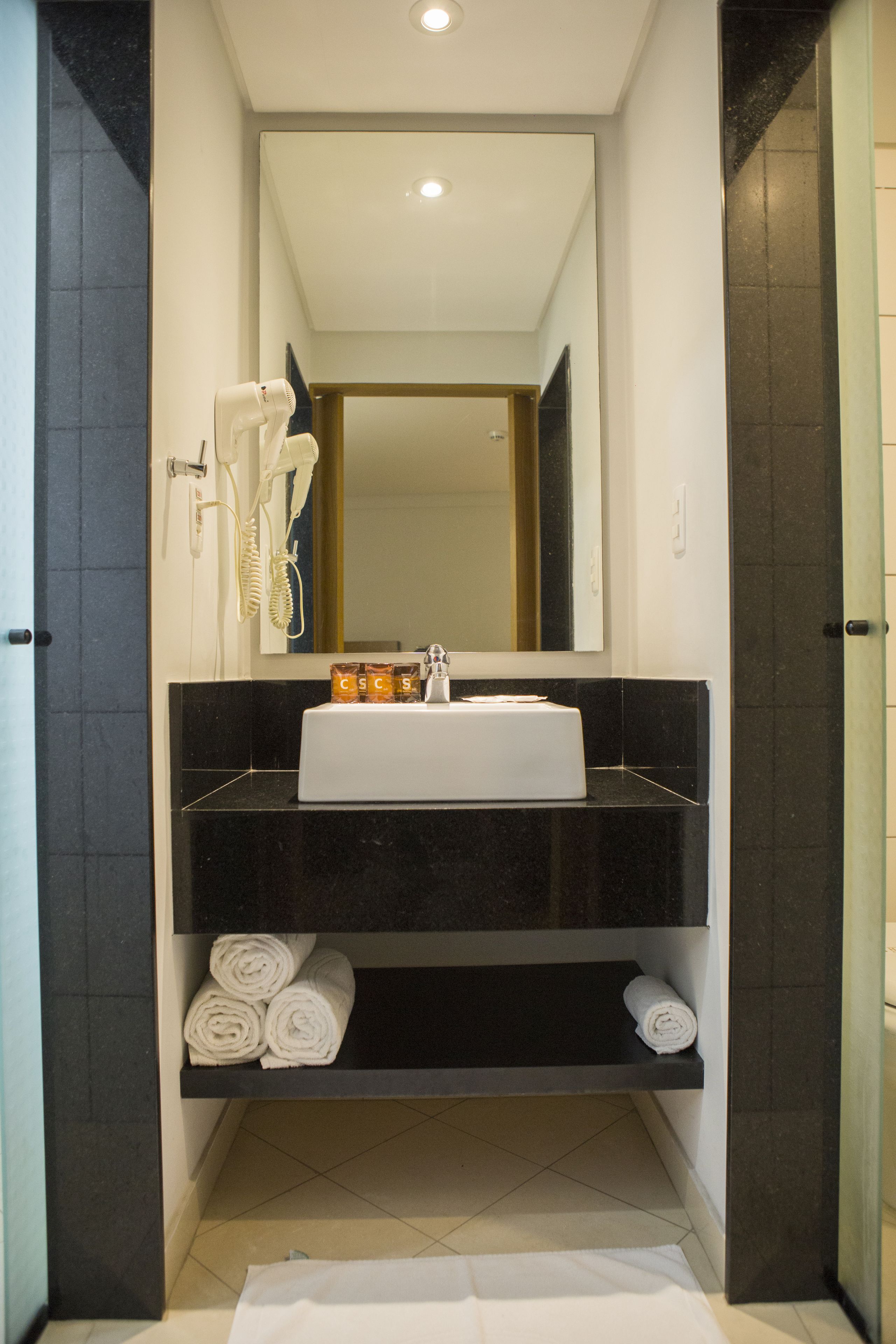 Een moderne badkamer met een zwarte aanrecht, een witte wastafel en een spiegel erboven. Handdoeken liggen op het onderste plankje en een haardroger zit aan de muur.