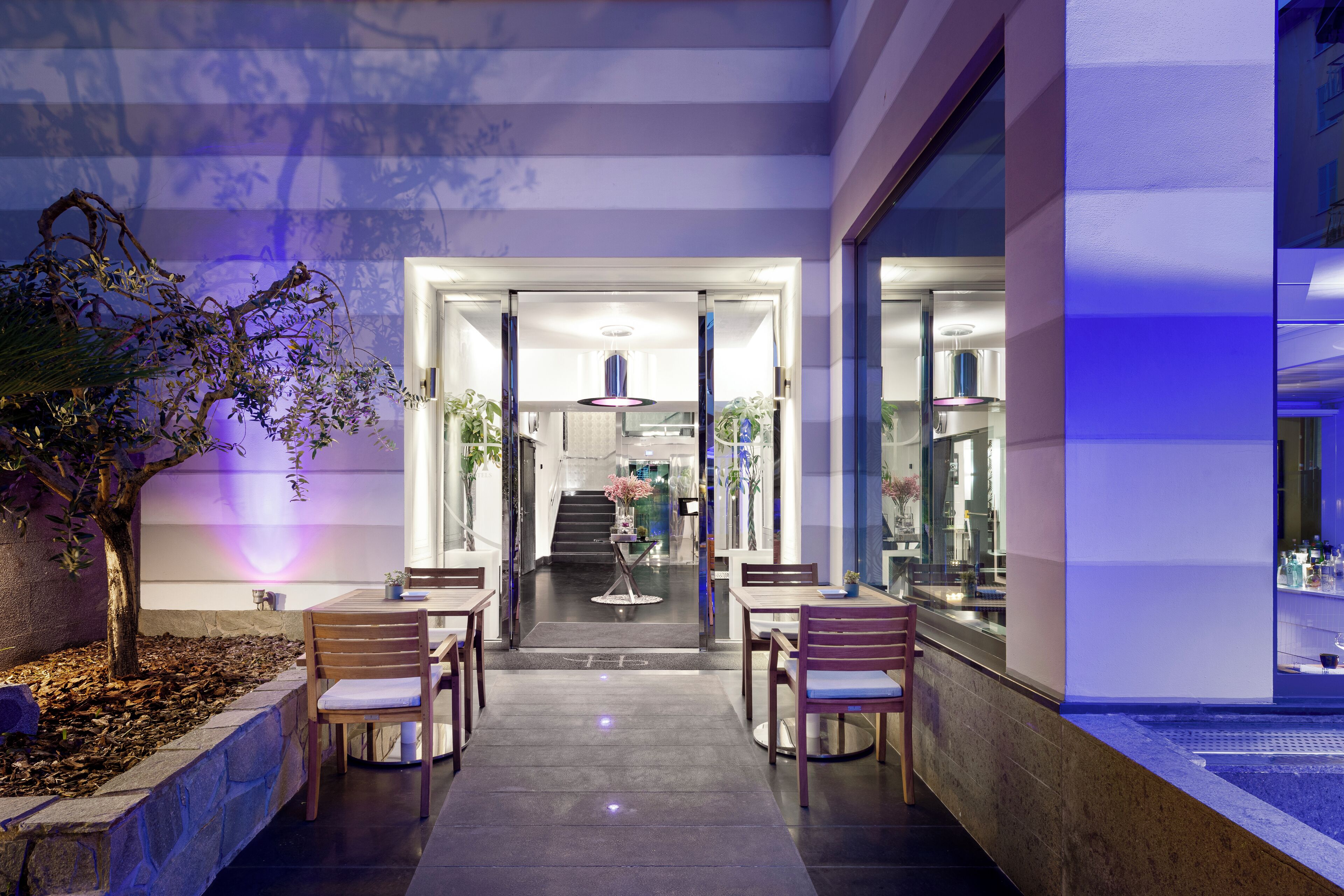 A modern hotel lobby with a glass entrance, wooden chairs, a central table with a vase, and a staircase leading to the upper floors.
