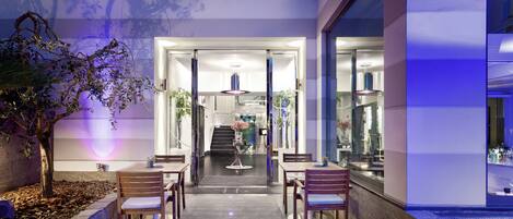 A modern hotel lobby with a glass entrance, wooden chairs, a central table with a vase, and a staircase leading to the upper floors.