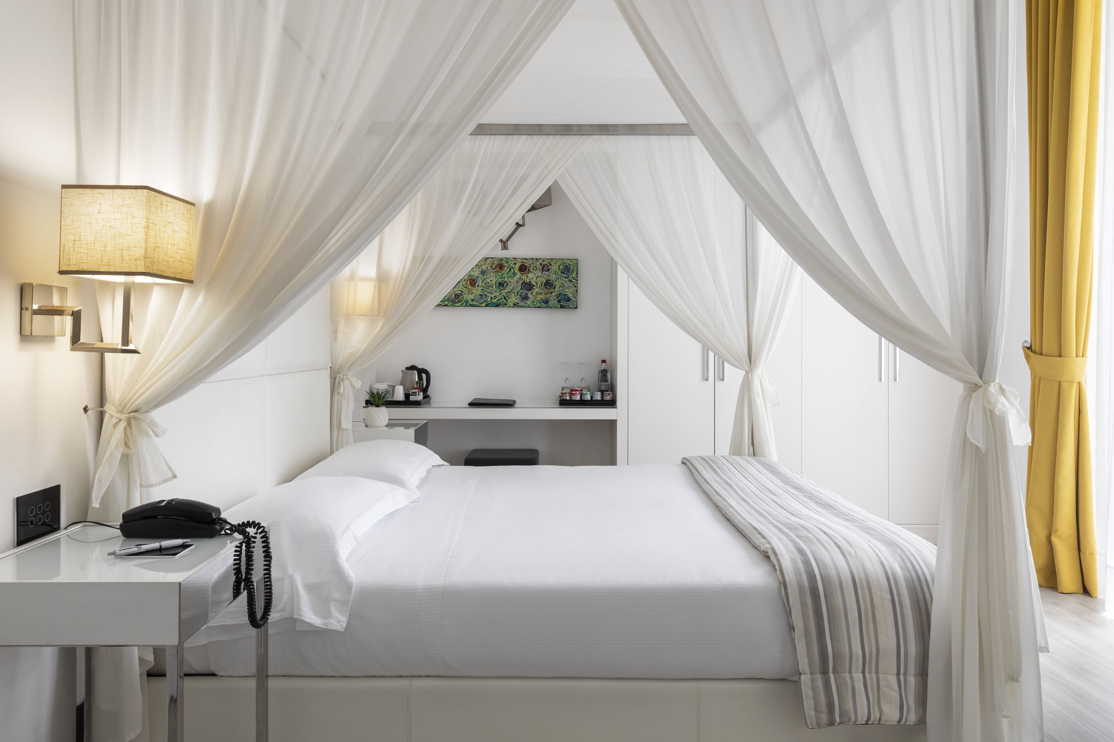 A hotel room with a bed, a desk with a lamp, a telephone, and a painting on the wall.