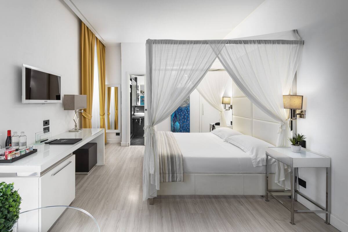 A modern hotel room with a bed, bedside table, TV, and a desk with a computer.