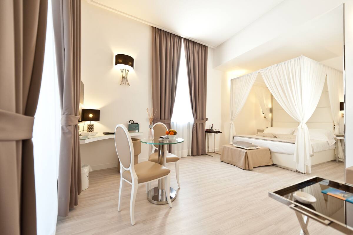A hotel room with a bed, a glass table, a chair, a desk, and curtains.