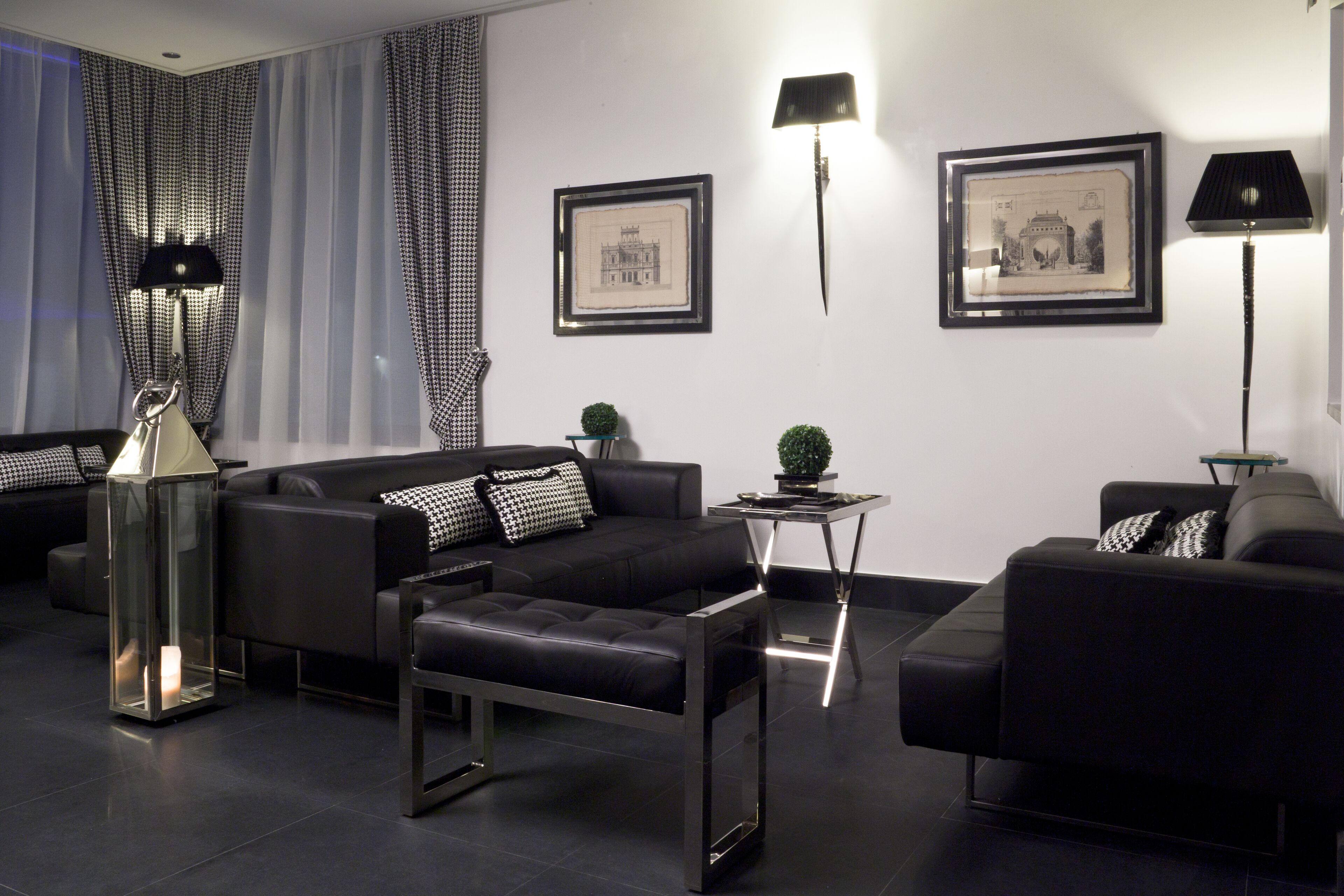 A modern living room with black sofas, a glass coffee table, and black and white patterned cushions. There are framed pictures on the wall and two floor lamps.