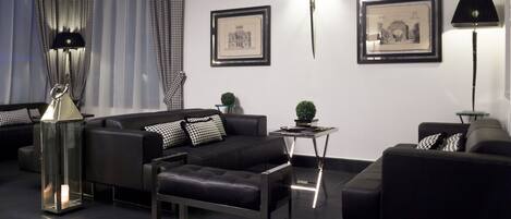 A modern living room with black sofas, a glass coffee table, and black and white patterned cushions. There are framed pictures on the wall and two floor lamps.
