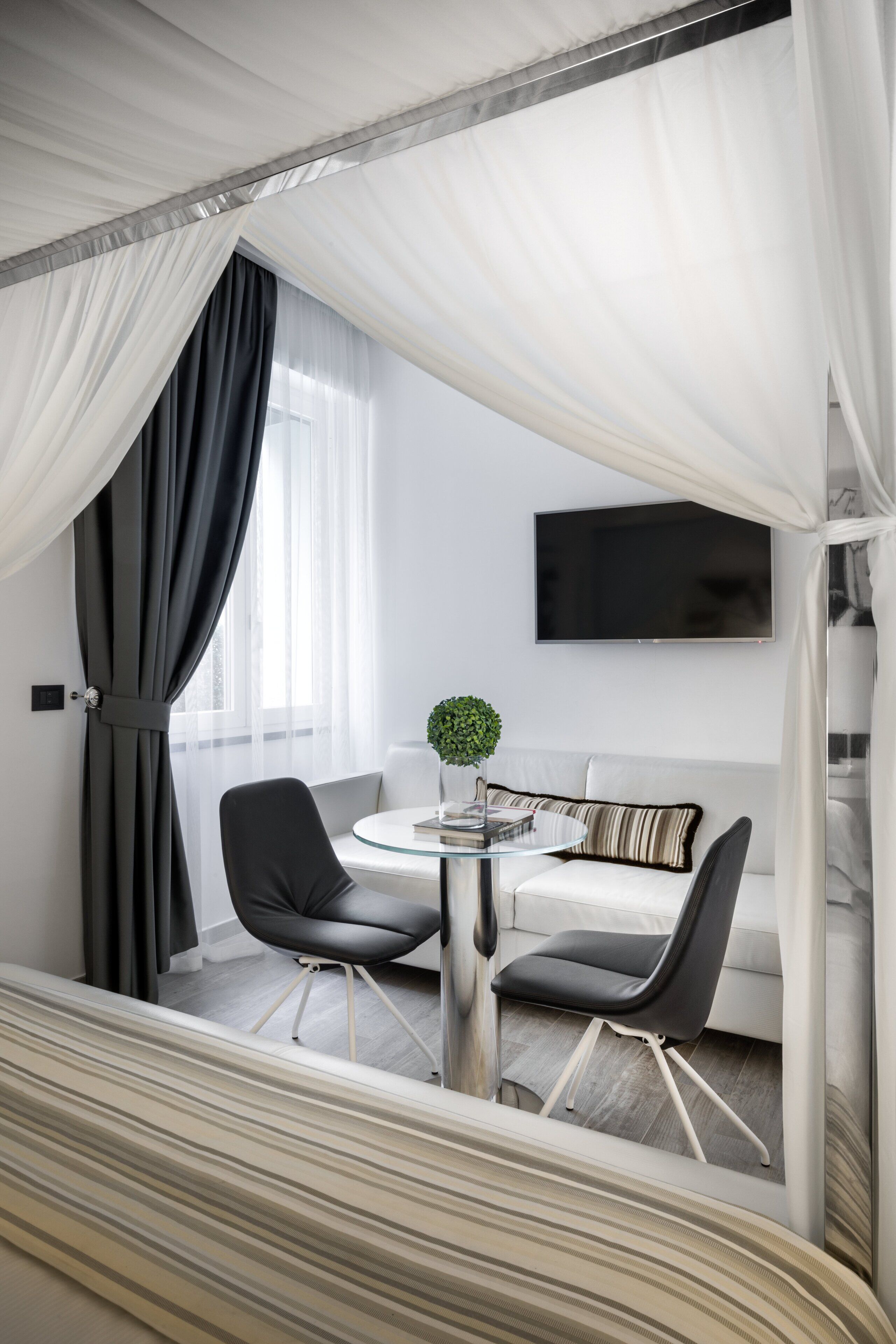 A modern hotel room with a bed, a round table with two chairs, a TV mounted on the wall, and a large window with sheer curtains.