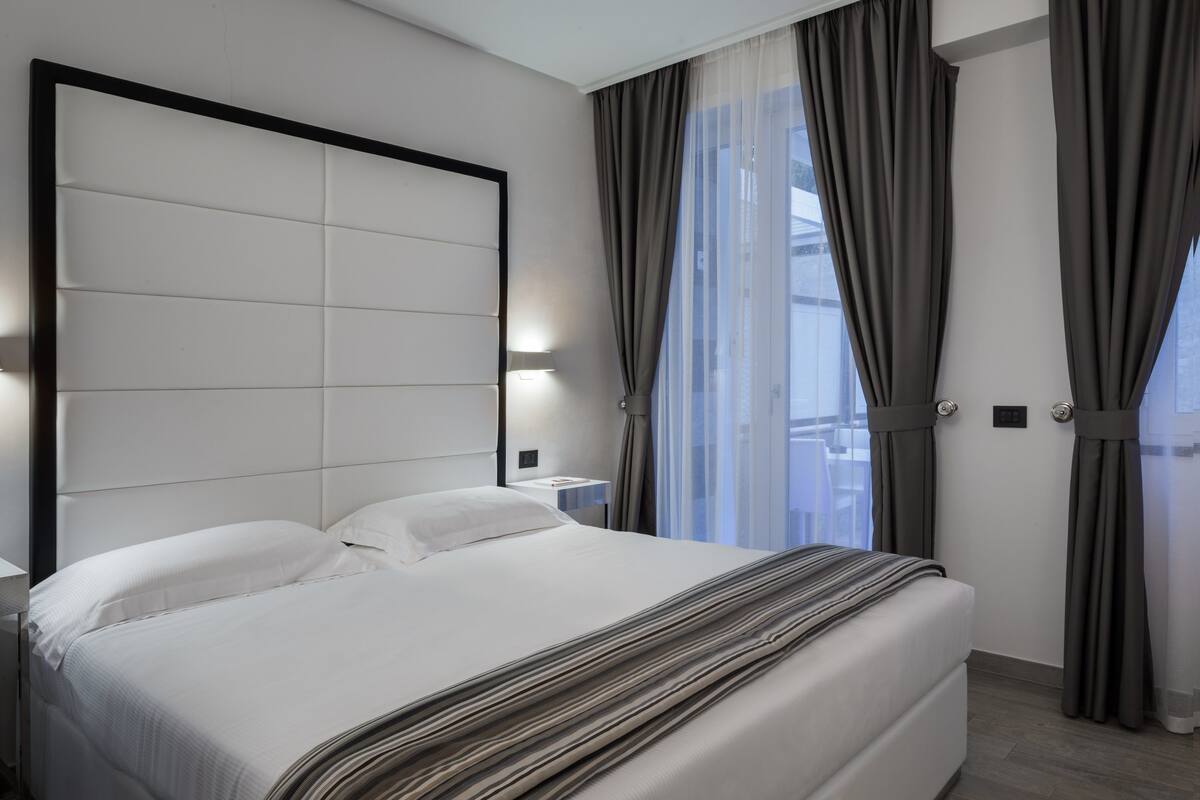 A modern hotel room with a large bed, a headboard, a bedside table, and a view of the city through sheer curtains.