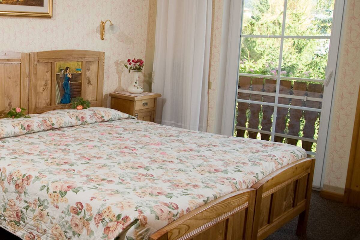 A bedroom with a wooden bed, a floral chair, a nightstand with a vase of flowers, and a window with a view of greenery.