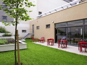 Une cour avec un petit arbre, des chaises et des tables rouges, un escalier et un bâtiment doté de grandes fenêtres.