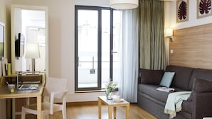 A modern living room with a sofa, a desk with a laptop, a chair, a small table with a plant, and a large window with curtains.