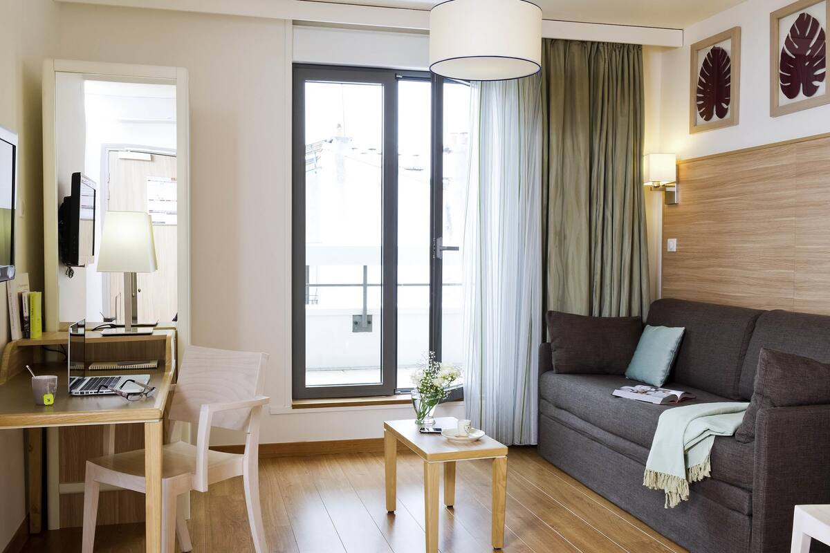 Un salon moderne comprenant un canapé, un bureau avec un ordinateur portable, une chaise, une petite table avec une plante et une grande fenêtre avec des rideaux.