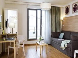 Uma sala de estar moderna com um sofá, uma escrivaninha com um laptop, uma cadeira, uma pequena mesa com uma planta e uma grande janela com cortinas.