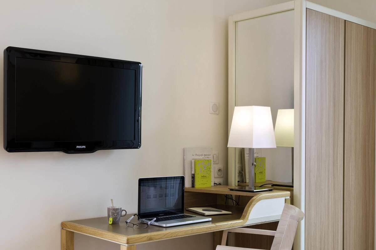 Ein Hotelzimmer mit Flachbildfernseher, einem Holztisch mit Laptop, einem Sessel und einer Nachttischlampe.