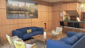 A modern hotel lobby with blue sofas, a wooden wall with a large lake painting, a small bar area, and a fire extinguisher.