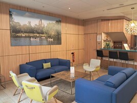 Um hall de hotel moderno com sofás azuis, uma parede de madeira com uma grande pintura de um lago, uma pequena área de bar e um extintor de incêndio.