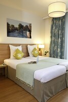 Quarto de hotel com cama, mesa de cabeceira, luminária, quadro e janela com cortinas.