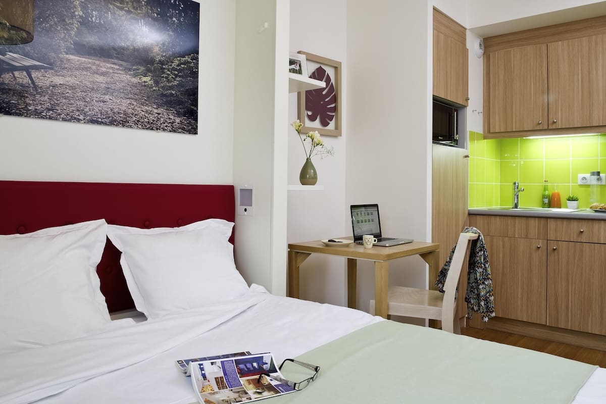 Une chambre d’hôtel comprenant un lit, un bureau avec un ordinateur portable, une petite table avec un vase, et un coin cuisine avec des placards en bois et un plan de travail vert.