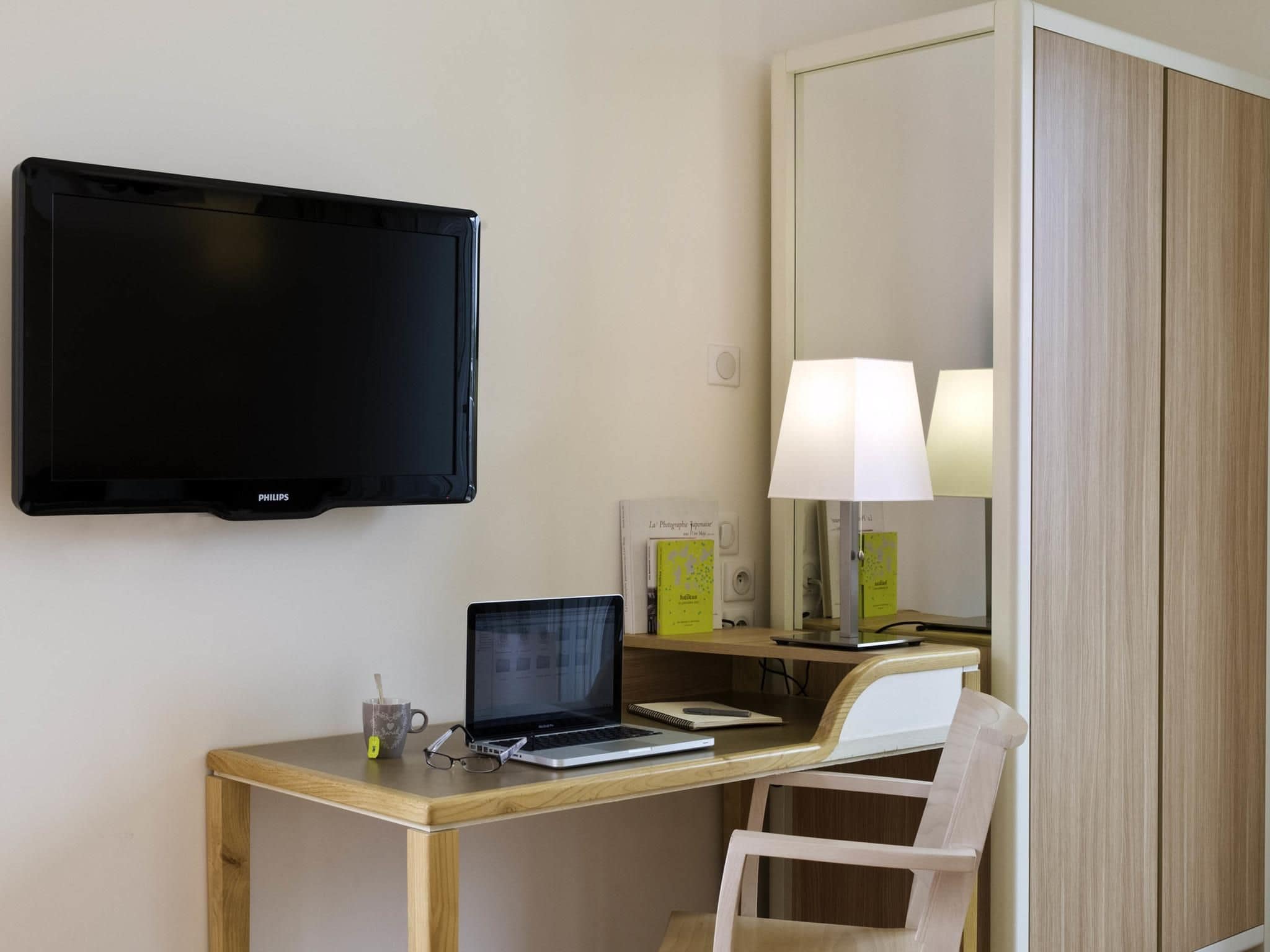 A hotel room with a flat-screen TV mounted on the wall, a desk with a laptop, a chair, a lamp, and a mirror.