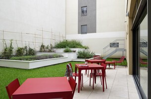 Um pátio com mesas e cadeiras vermelhas, um pequeno jardim com plantas e uma escadaria que leva a um andar superior.