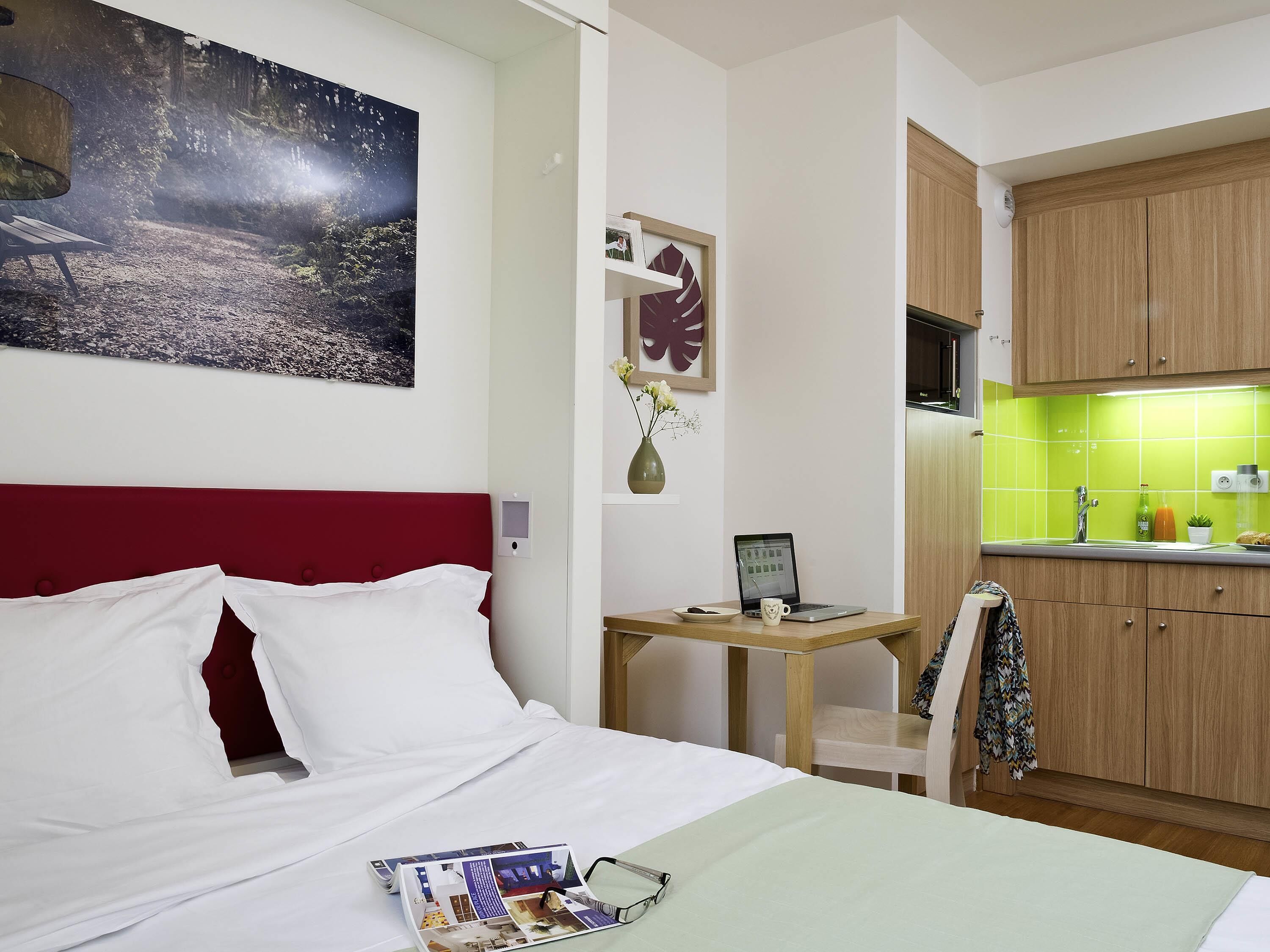 Een hotelkamer met een bed, een bureau met een laptop, een kleine tafel met een vaas en een kitchenette met houten kastjes.