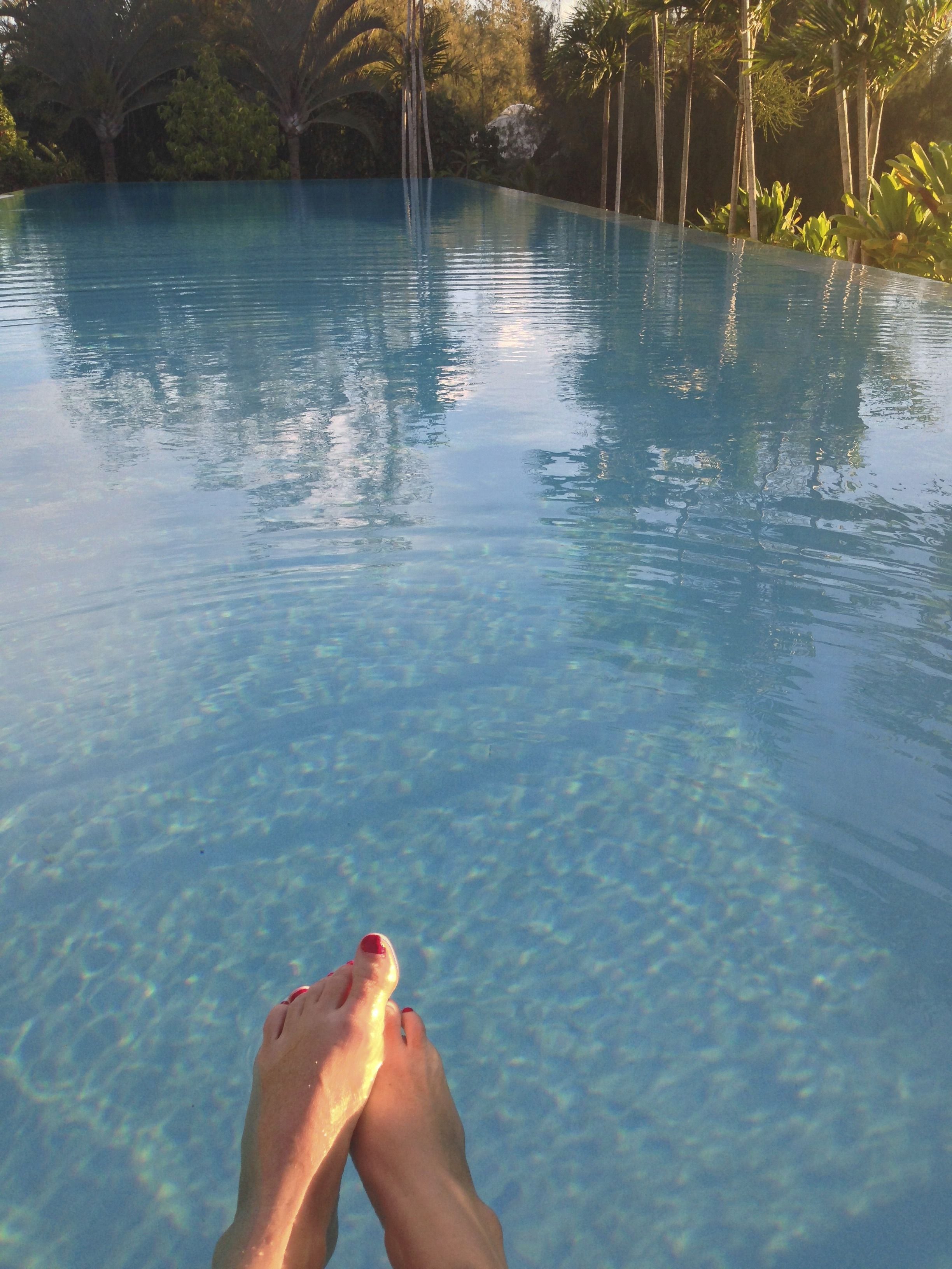 A clear view of a swimming pool with a person's feet at the edge, surrounded by lush greenery.