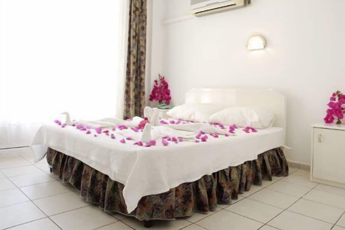Un lit bien fait, recouvert d’une courtepointe blanche et orné d’une jupe de lit à motifs floraux, une table de chevet blanche avec un vase de fleurs roses, et un mur blanc agrémenté d’un luminaire.