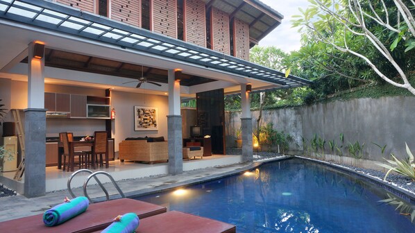 Un área de piscina moderna al aire libre, con una zona de estar cubierta, mobiliario para comer y un pequeño televisor.