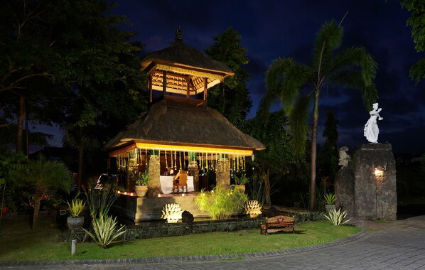 A well-lit outdoor bar area with a thatched roof, surrounded by tropical plants and a statue.