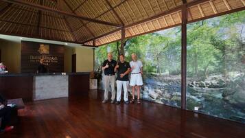 A reception area with a wooden reception desk, a large window with a view of a forest stream, and a thatched roof.