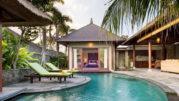 A swimming pool with lounge chairs, a thatched-roof structure, and a view of a building with a tiled roof.