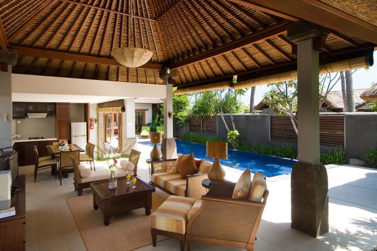 Un área de estar al aire libre, techada, con piscina, sillones de descanso y una mesa de comedor.