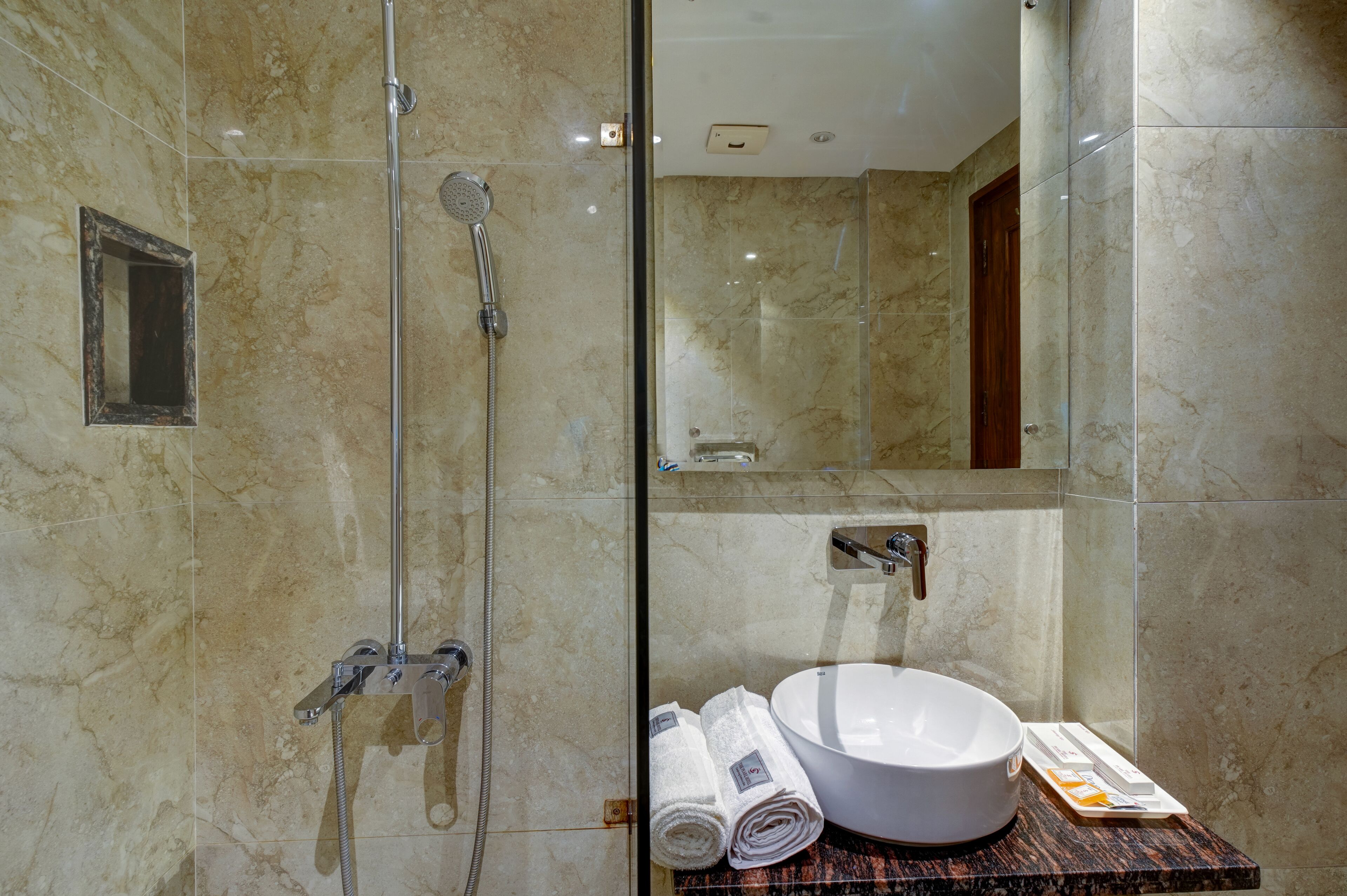 A modern bathroom with a glass-enclosed shower, a marble-tiled wall, a sink with a large basin, and neatly rolled towels on the counter.