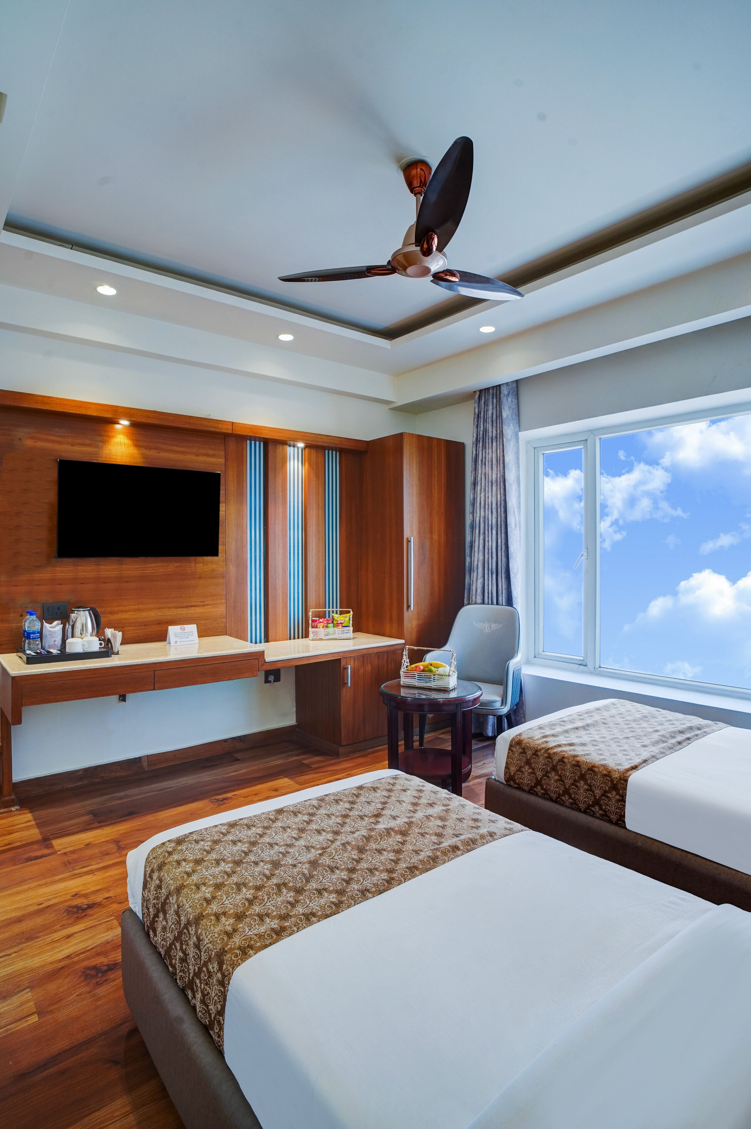 A hotel room with two beds, a flat-screen TV mounted on the wall, a ceiling fan, a window with a view of the sky, and a small table with a chair.
