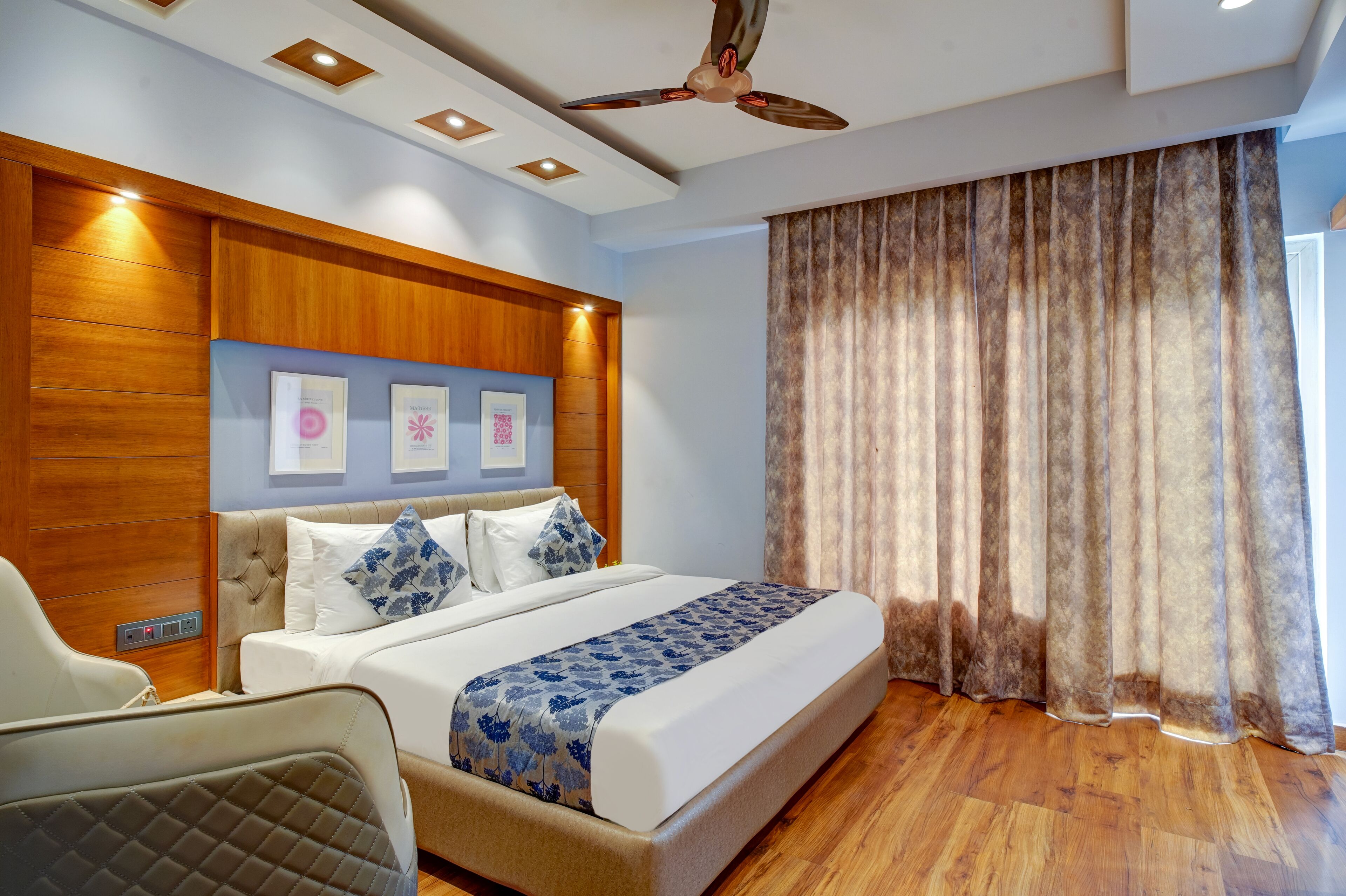 A hotel room with a bed, a headboard with framed pictures, a ceiling fan, and a large window with curtains.