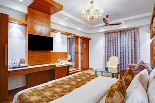 A hotel room with a large bed, a desk with a flat-screen TV, a wardrobe, a seating area with a chair, and a ceiling fan.