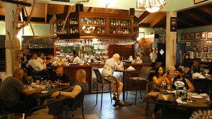 A bar area with wooden shelves stocked with bottles, a bar counter, and patrons seated at tables.
