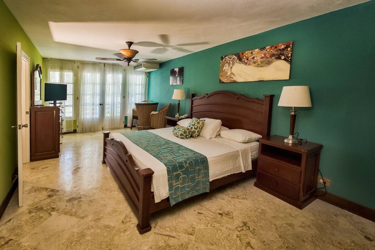 A bedroom with a bed, nightstands, a desk, a chair, a ceiling fan, and a painting on the wall.
