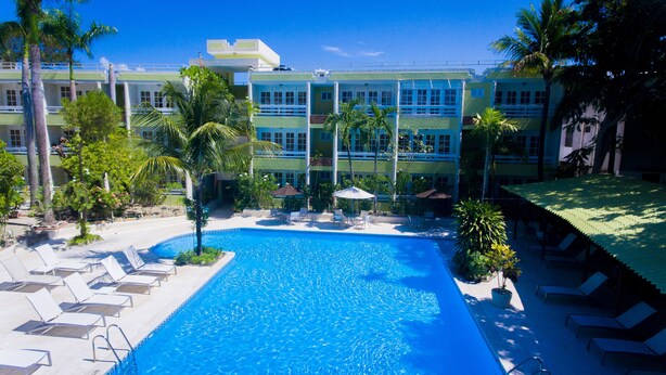 A swimming pool with lounge chairs and palm trees, surrounded by a multi-story building with balconies.