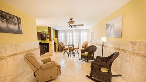 A living room with a beige armchair, a wicker rocking chair, a dining table with chairs, a ceiling fan, and artwork on the walls.