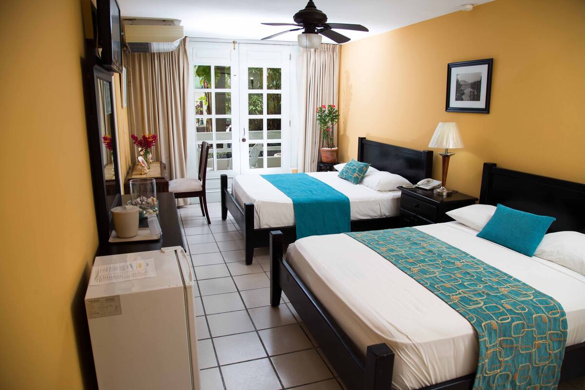 A hotel room with two beds, a ceiling fan, a potted plant, a desk with a chair, and a large window with curtains.