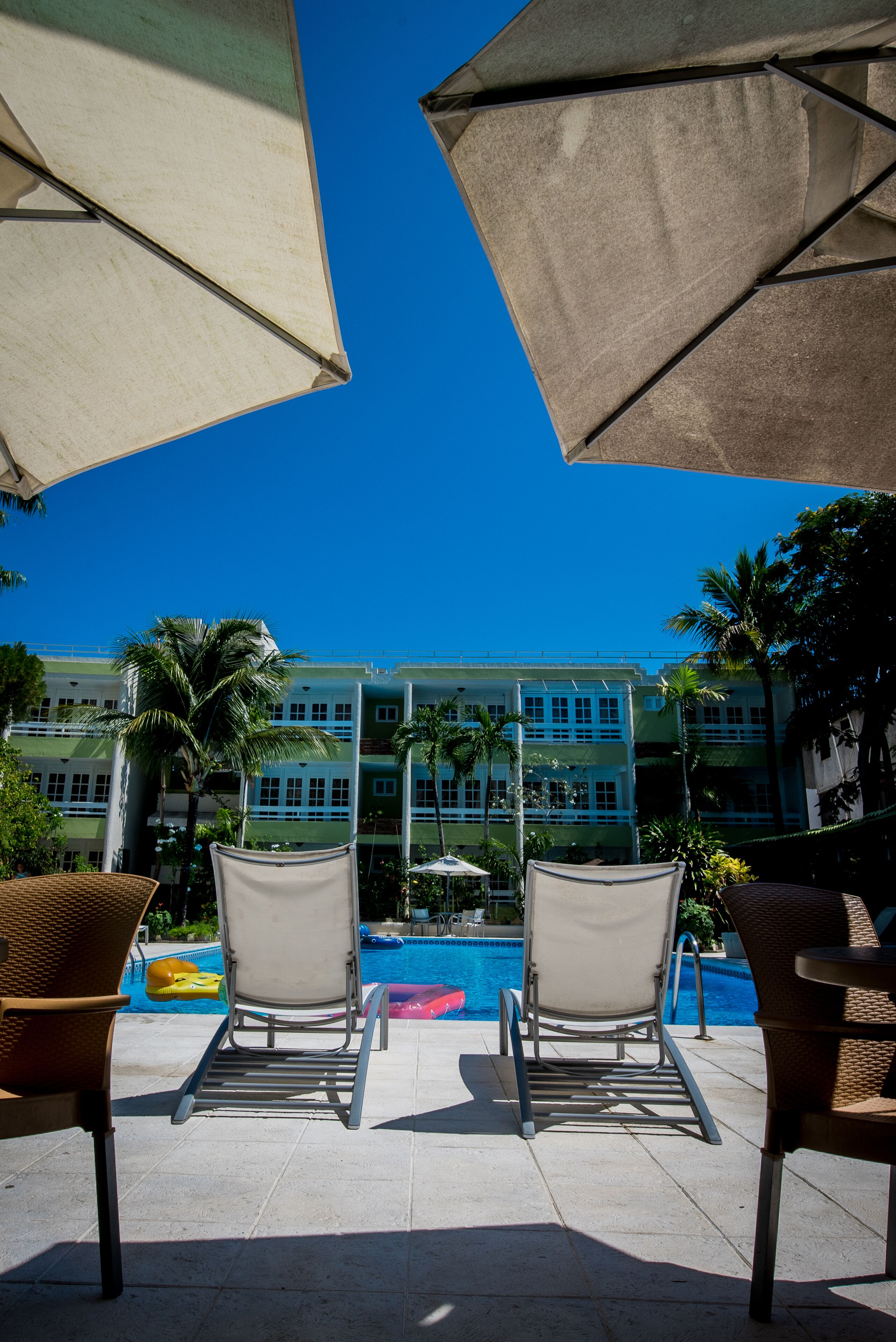 A poolside area with lounge chairs and umbrellas, a clear blue sky, and a building with multiple floors in the background.