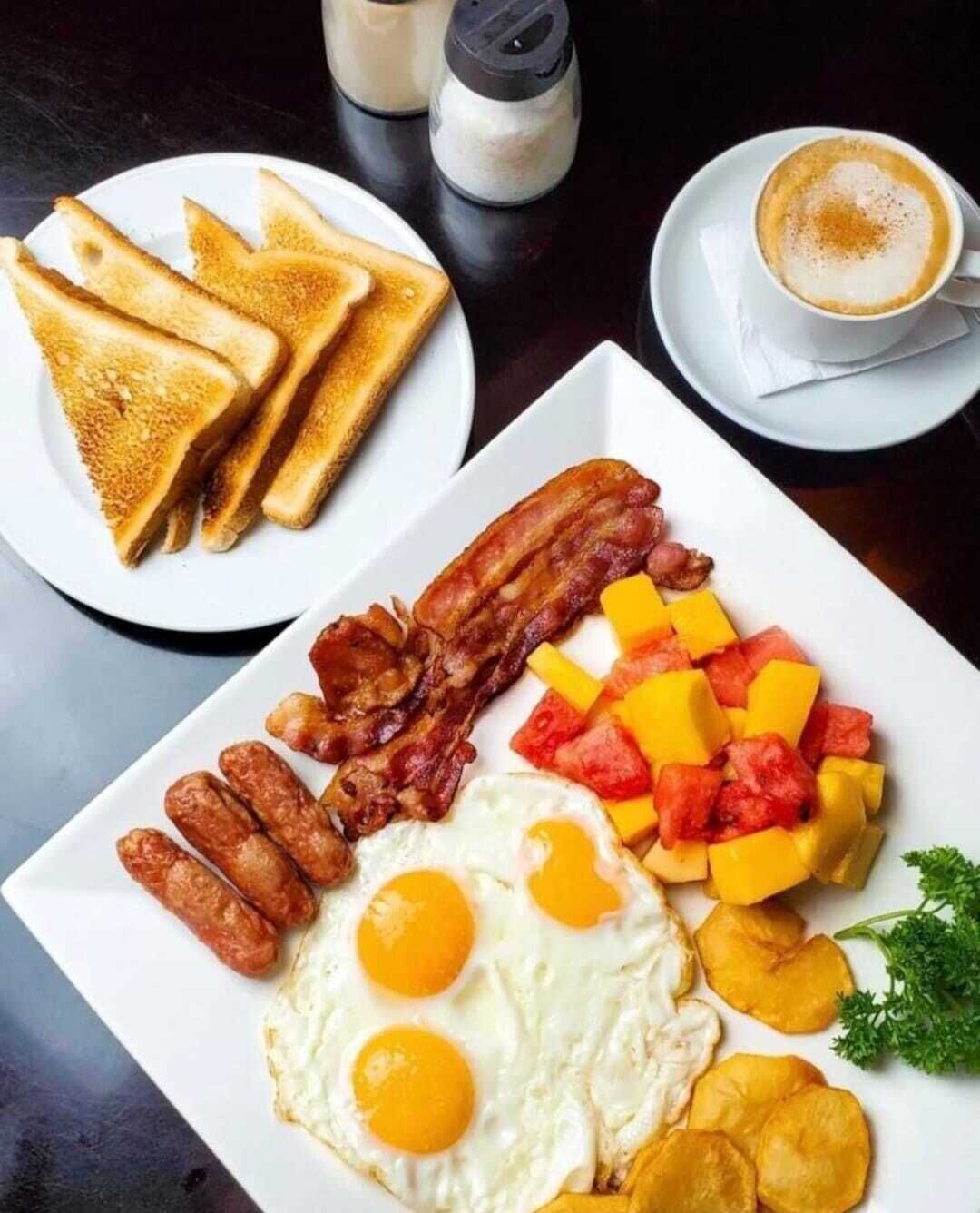 A breakfast plate with toast, bacon, sausages, eggs, and fruit.