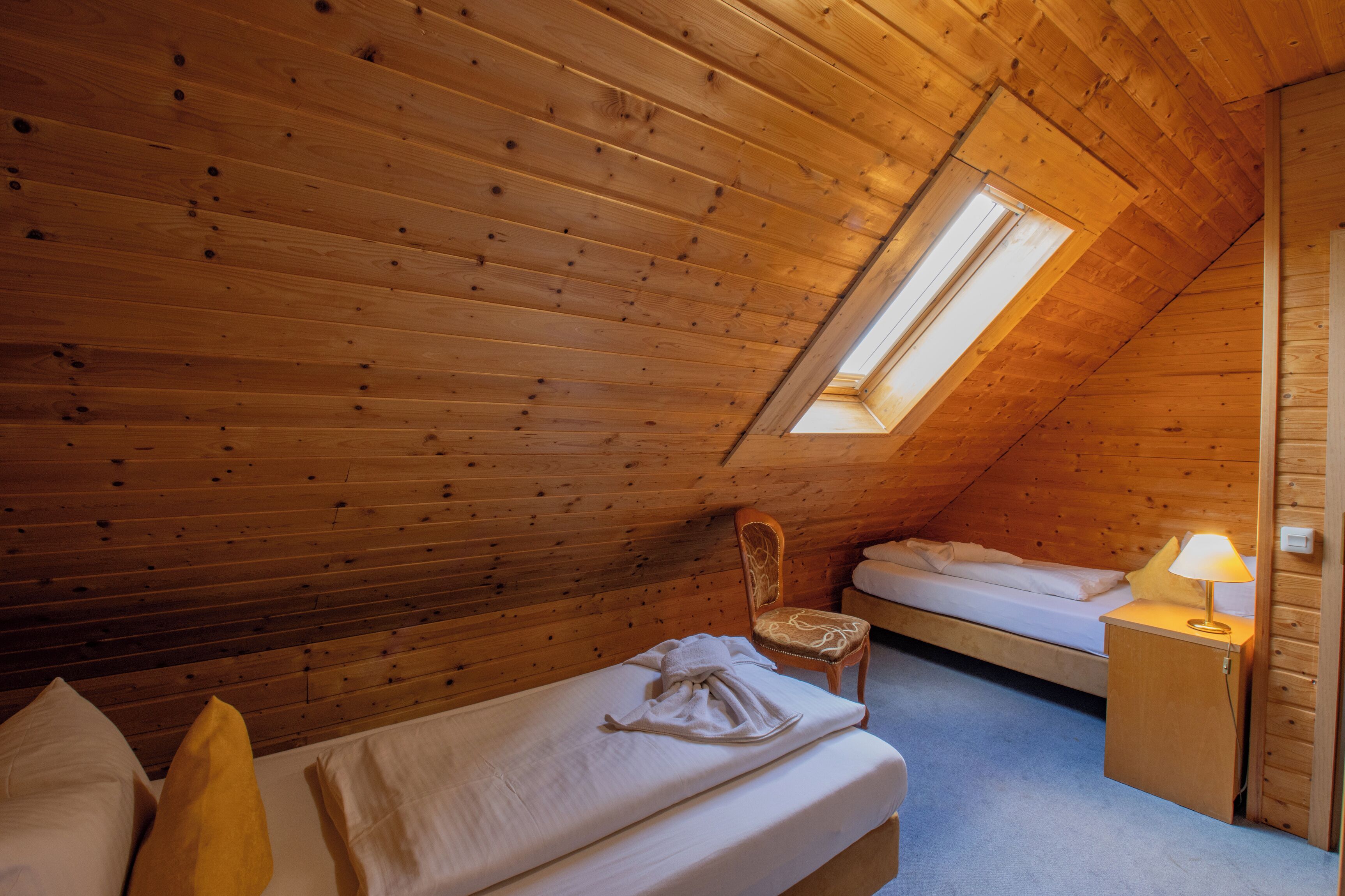 A room with a sloped ceiling, wooden walls, a single bed, a chair, a bedside table with a lamp, and a window.