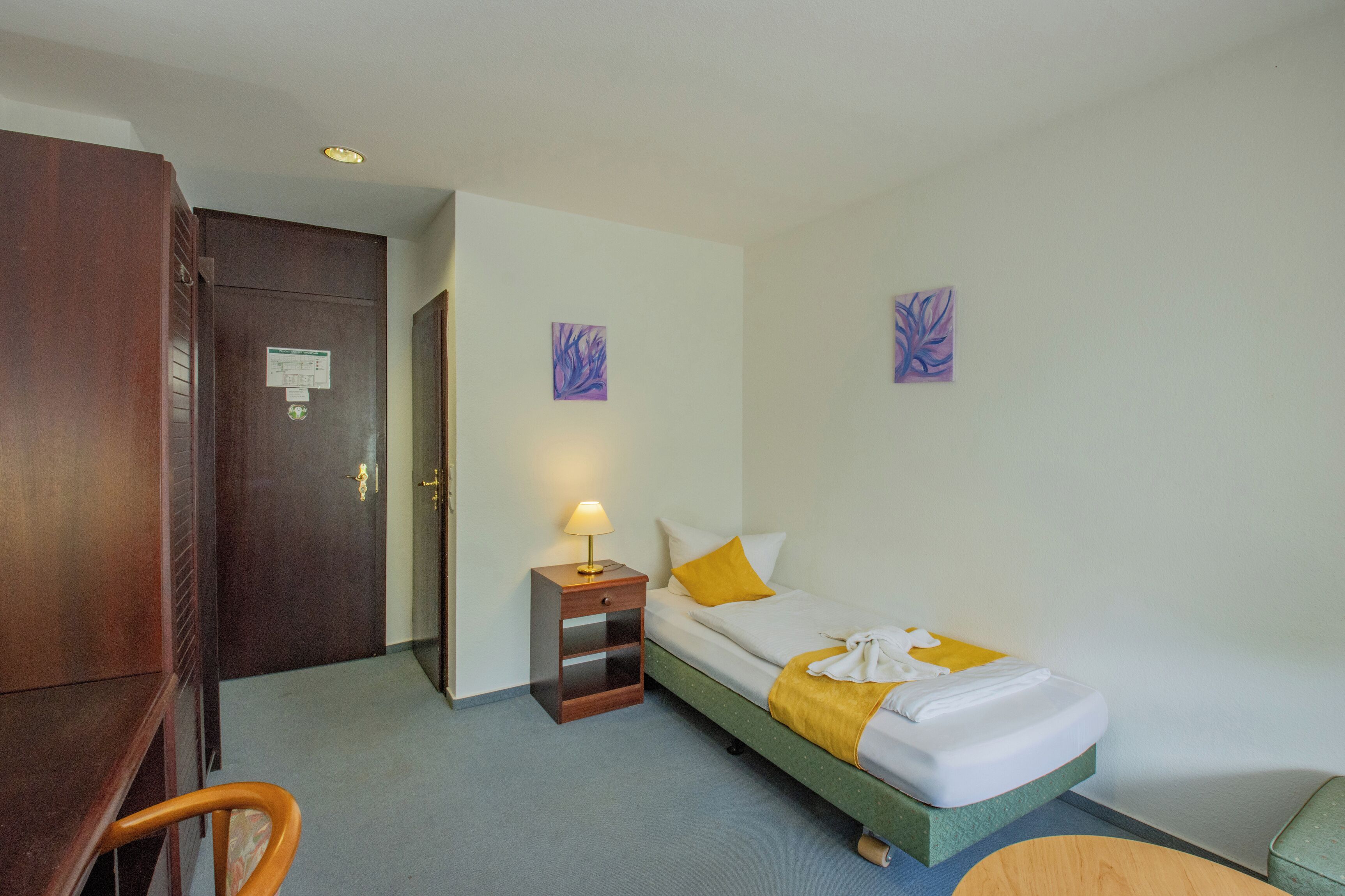 A hotel room with a bed, a nightstand, a chair, and two wall-mounted paintings.