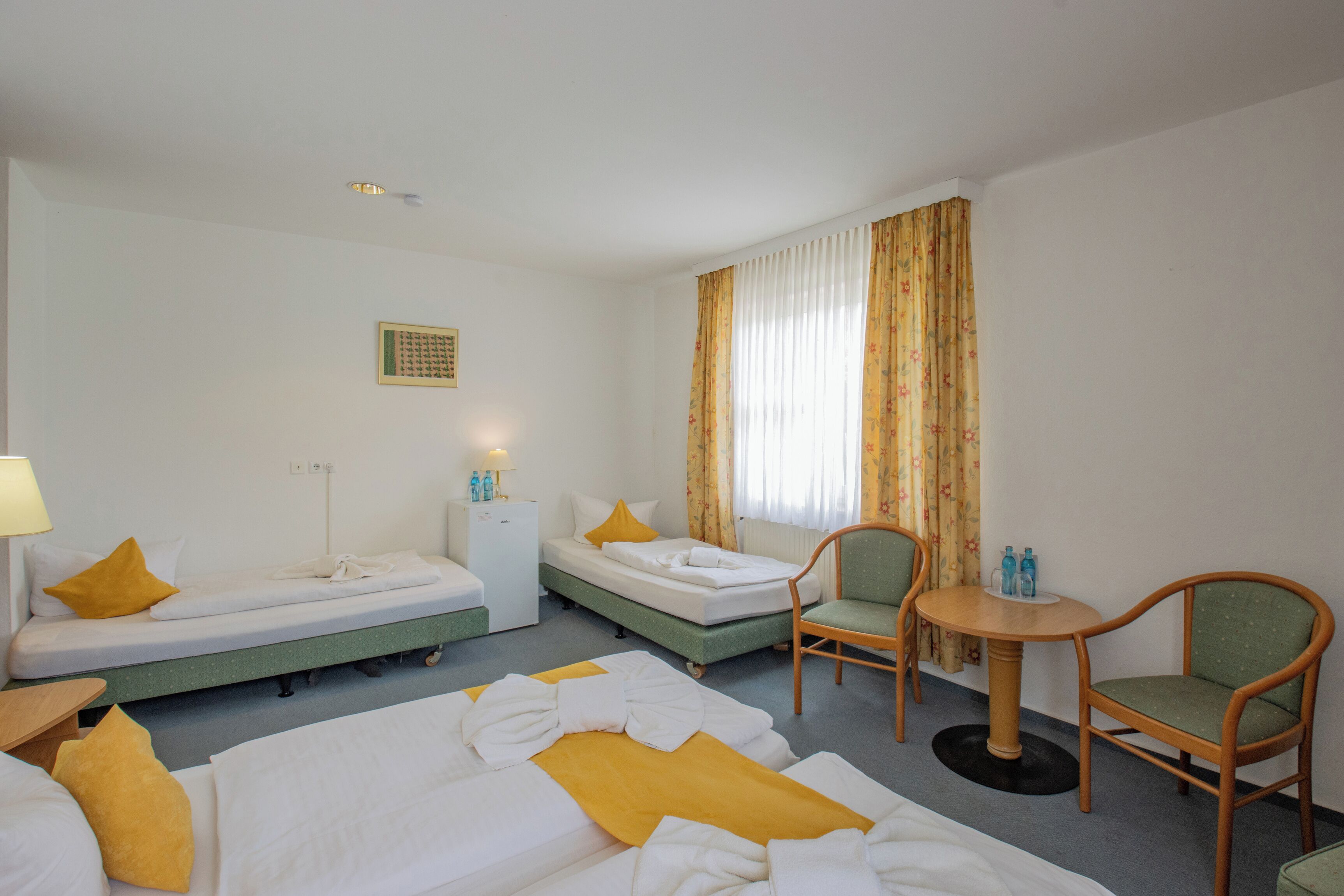 A hotel room with two single beds, a small table with chairs, a refrigerator, and a window with curtains.