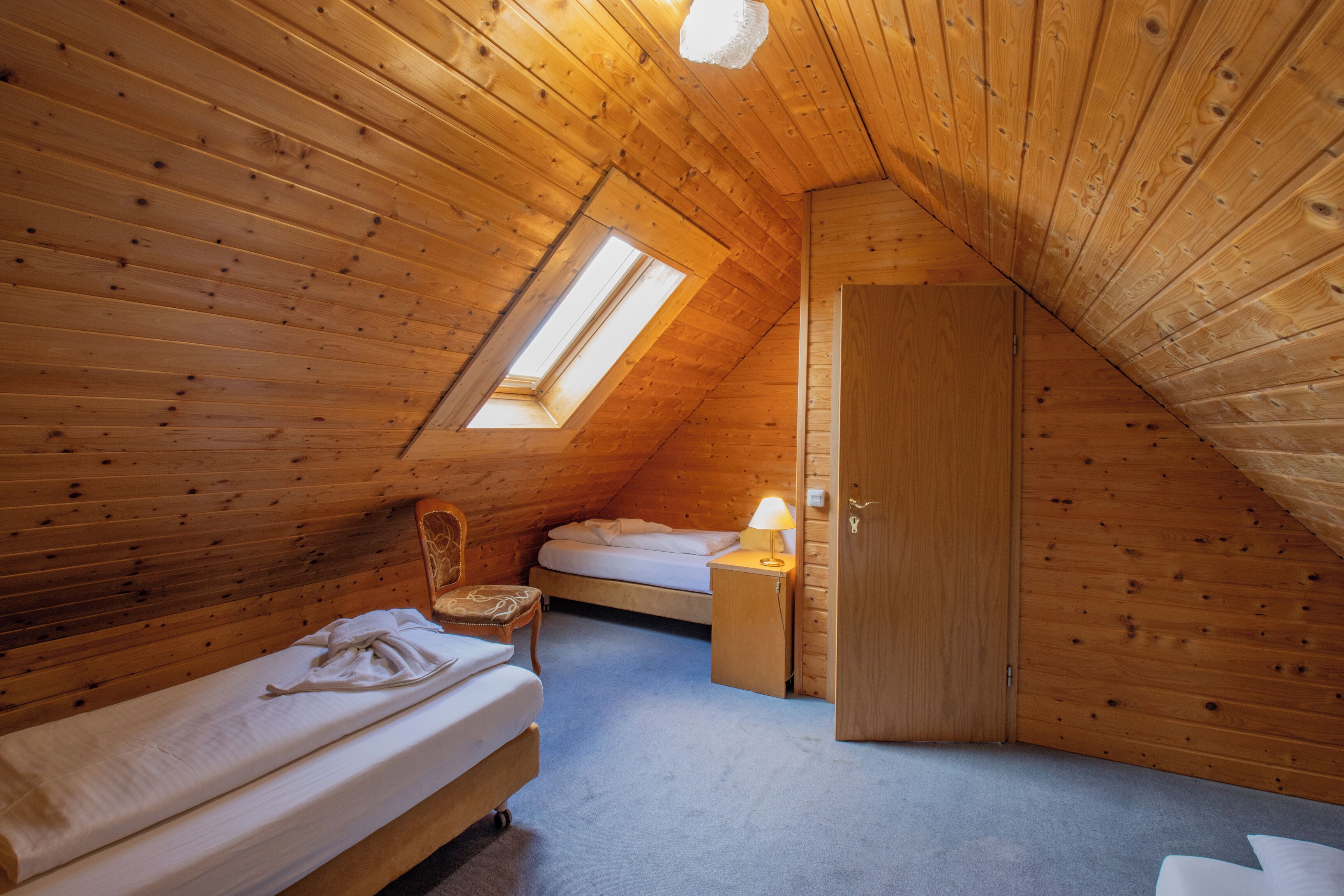 Ein Zimmer mit Holzboden und schrägem Dach, ausgestattet mit einem Bett, einem Sessel, einem Nachttisch mit Lampe und einer Tür.