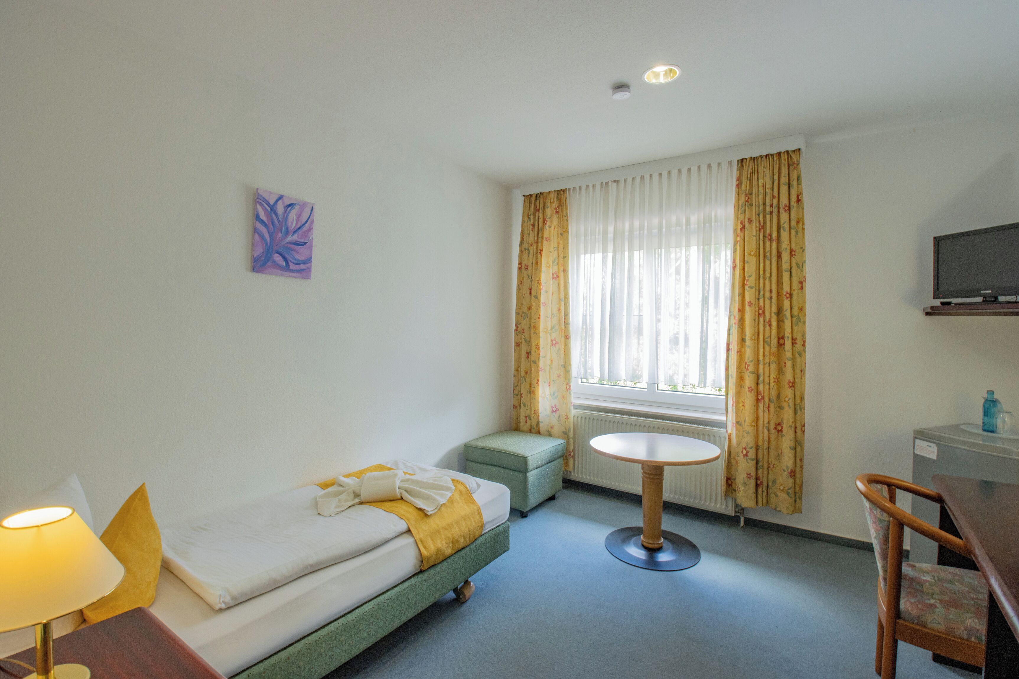 Habitación de hotel con cama, una mesita, una silla, televisor y ventana con cortinas.