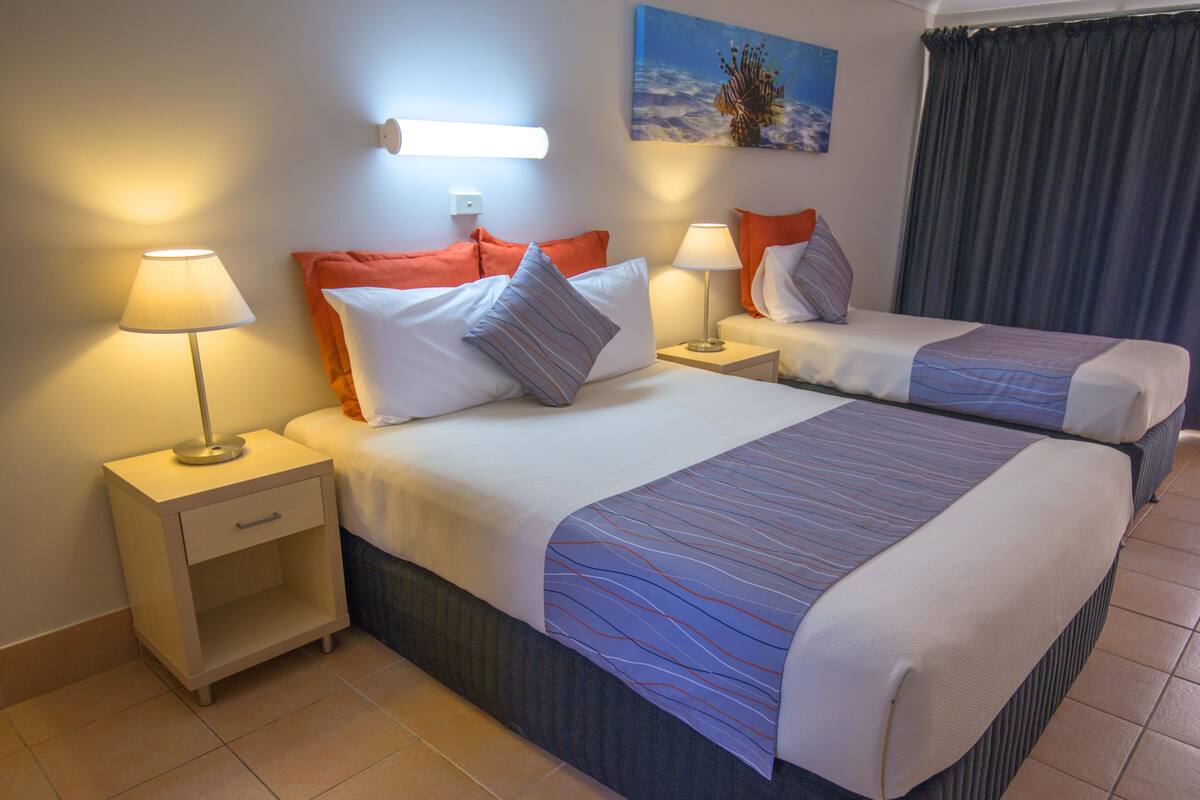 Habitación de hotel con dos camas, cada una con mesita de noche, lámparas y almohadas. Hay un cuadro en la pared y una cortina en la ventana.