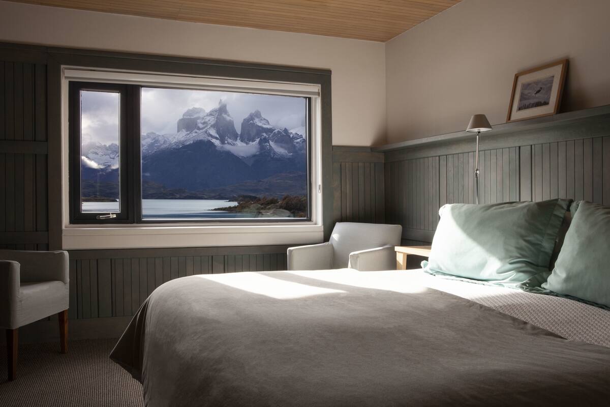 A bedroom with a large window, a bed with pillows, a chair, and a bedside table.